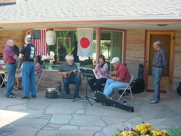 店には日本とアメリカの国旗が飾られ店の入口では男性がミニギターを女性がバイオリンを演奏しているのを横で眺めている男性1人と反対側では男性2人が話をしている様子の全体を映した写真